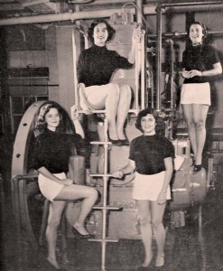 an black and white picture showing four women in matching outfits posing on a piece of engineering equipement
